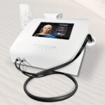 White cosmetic treatment device with a touchscreen showing a woman's face; has a handheld wand and cables on a sandy surface.